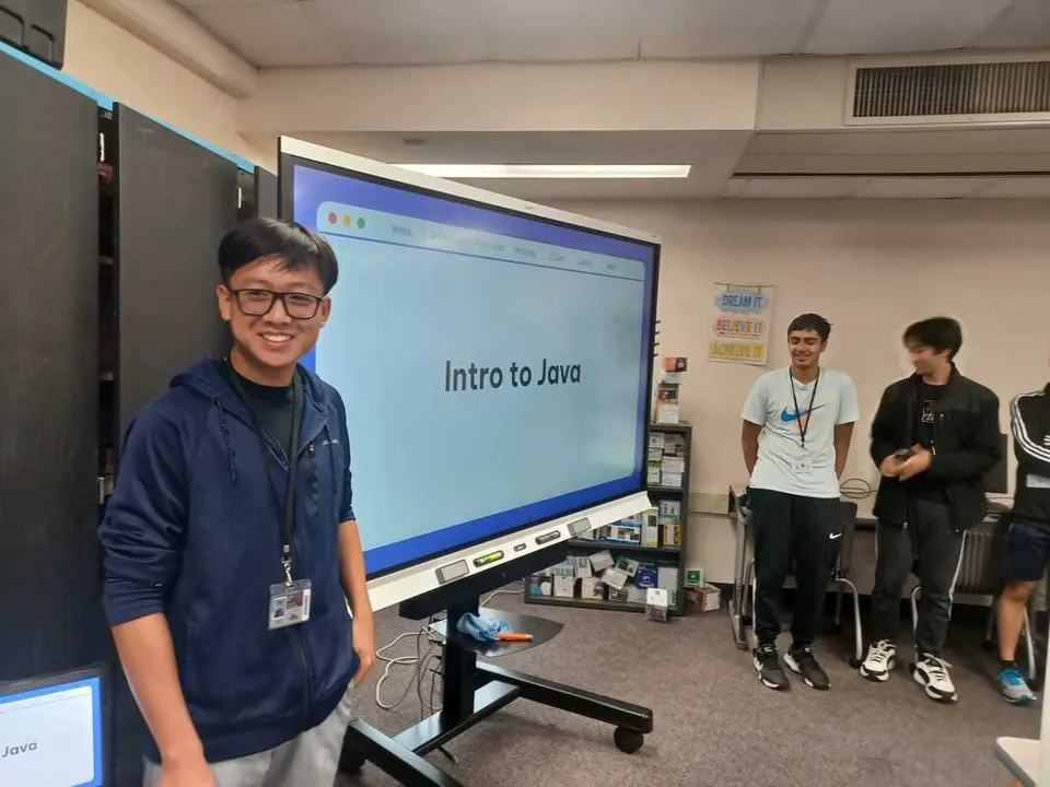 A member smiles next to the "Intro to Java" slide at a club meeting.